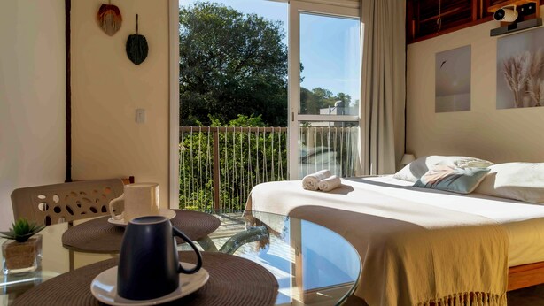 A modern hotel room with a balcony, a bed with white linens, a coffee table with a cup, and a view of greenery outside. - Pousada Contêiner 10 (Serra)