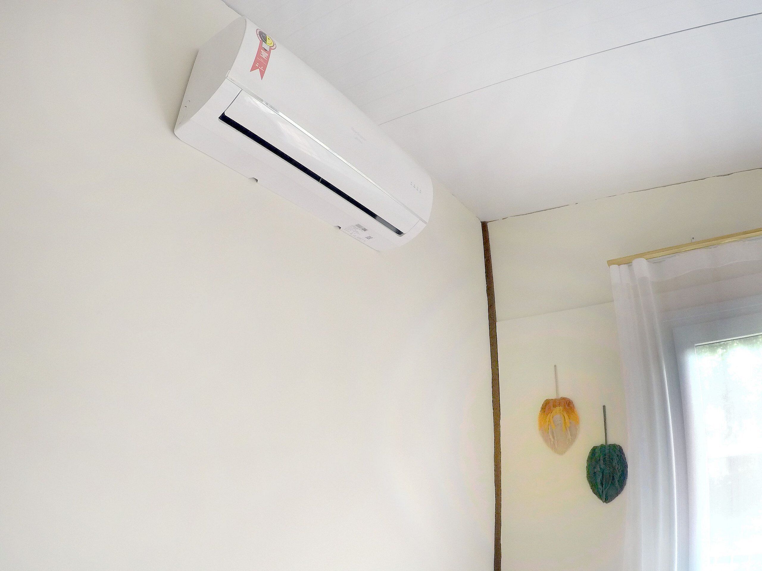 A room with a white ceiling, a white air conditioning unit, a window with white curtains, and a wall-mounted decorative item.