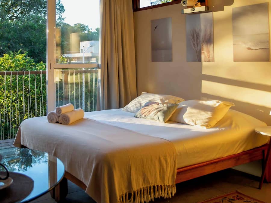 Een slaapkamer met een bed, kussens, een nachtkastje met een lamp en uitzicht op groen door een raam.