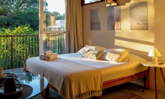 Un dormitorio con cama, almohadas, mesita de noche con lámpara y vista a zonas verdes por la ventana.