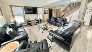 A spacious living room with a large flat-screen TV, leather sofas, a coffee table, and a staircase.