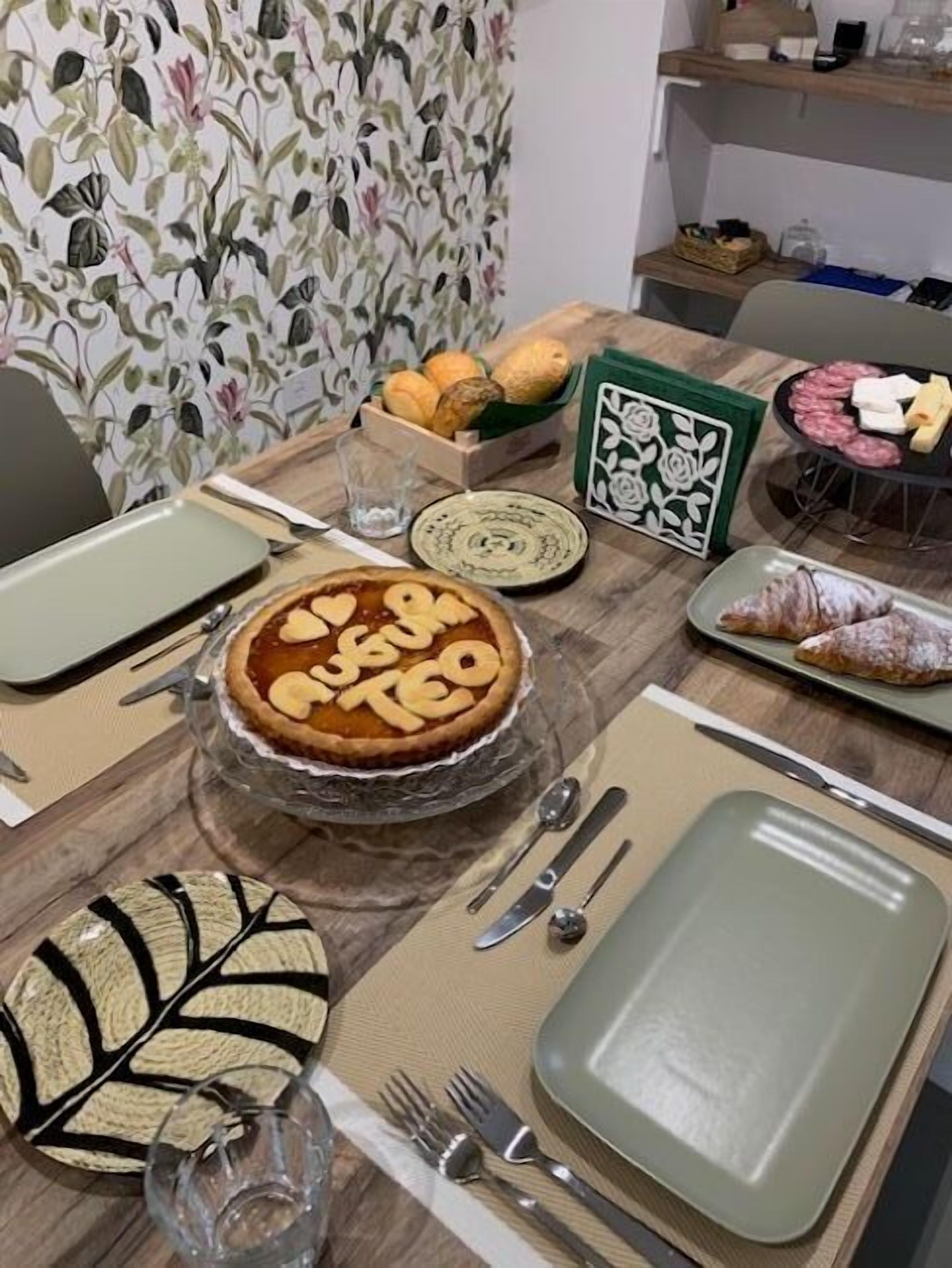 Una mesa de comedor dispuesta con platos, un pastel, un croissant y una hoja decorativa.