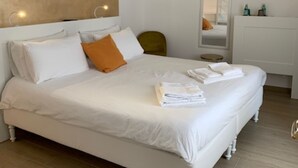 Een net opgemaakt bed met witte lakens en een oranje kussen, een witte dressoir met toiletartikelen, en een wandairconditioner.