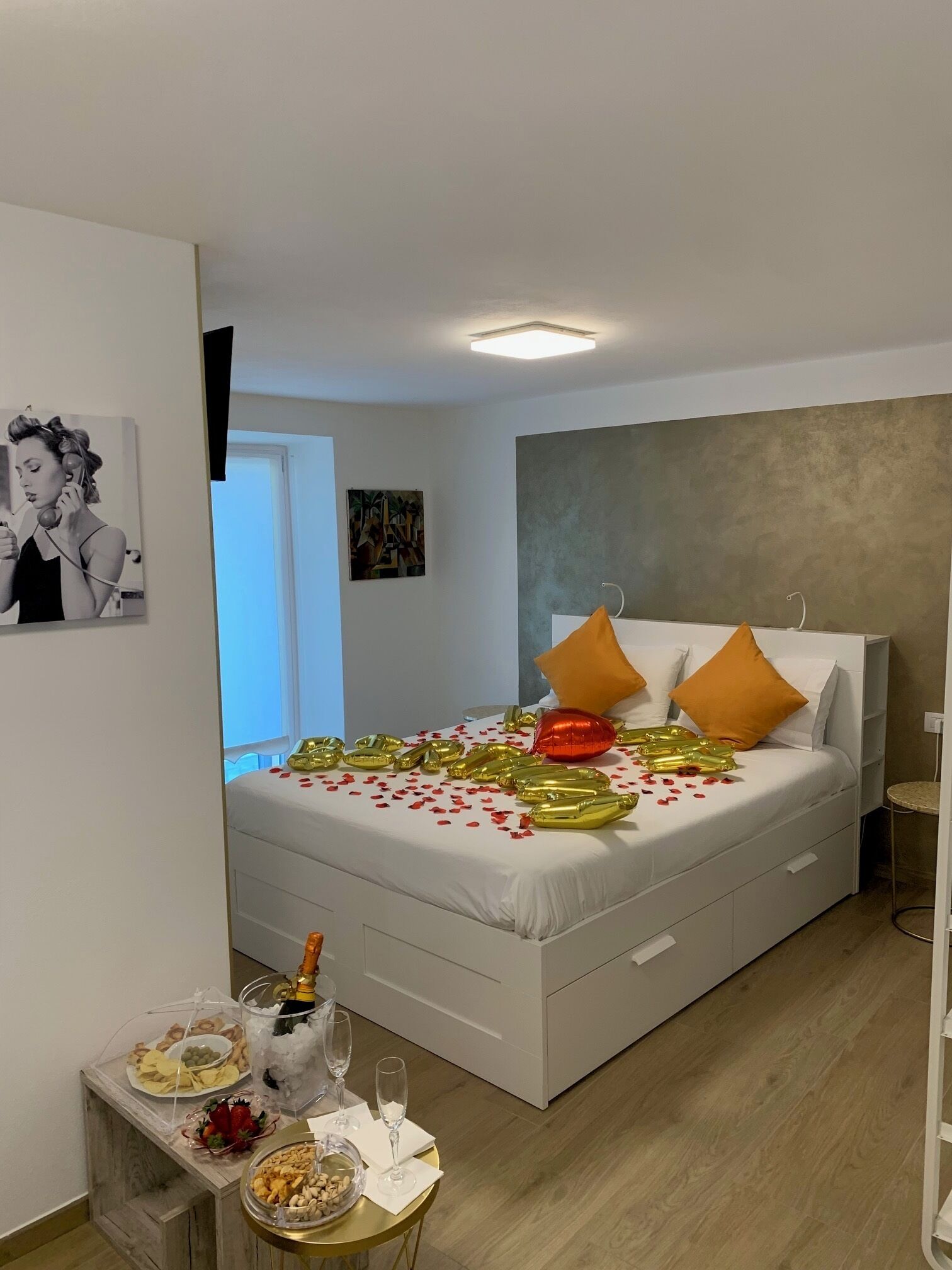 A modern bedroom with a bed, white bedding, and decorative pillows. There is a small table with snacks and drinks, and a picture on the wall.