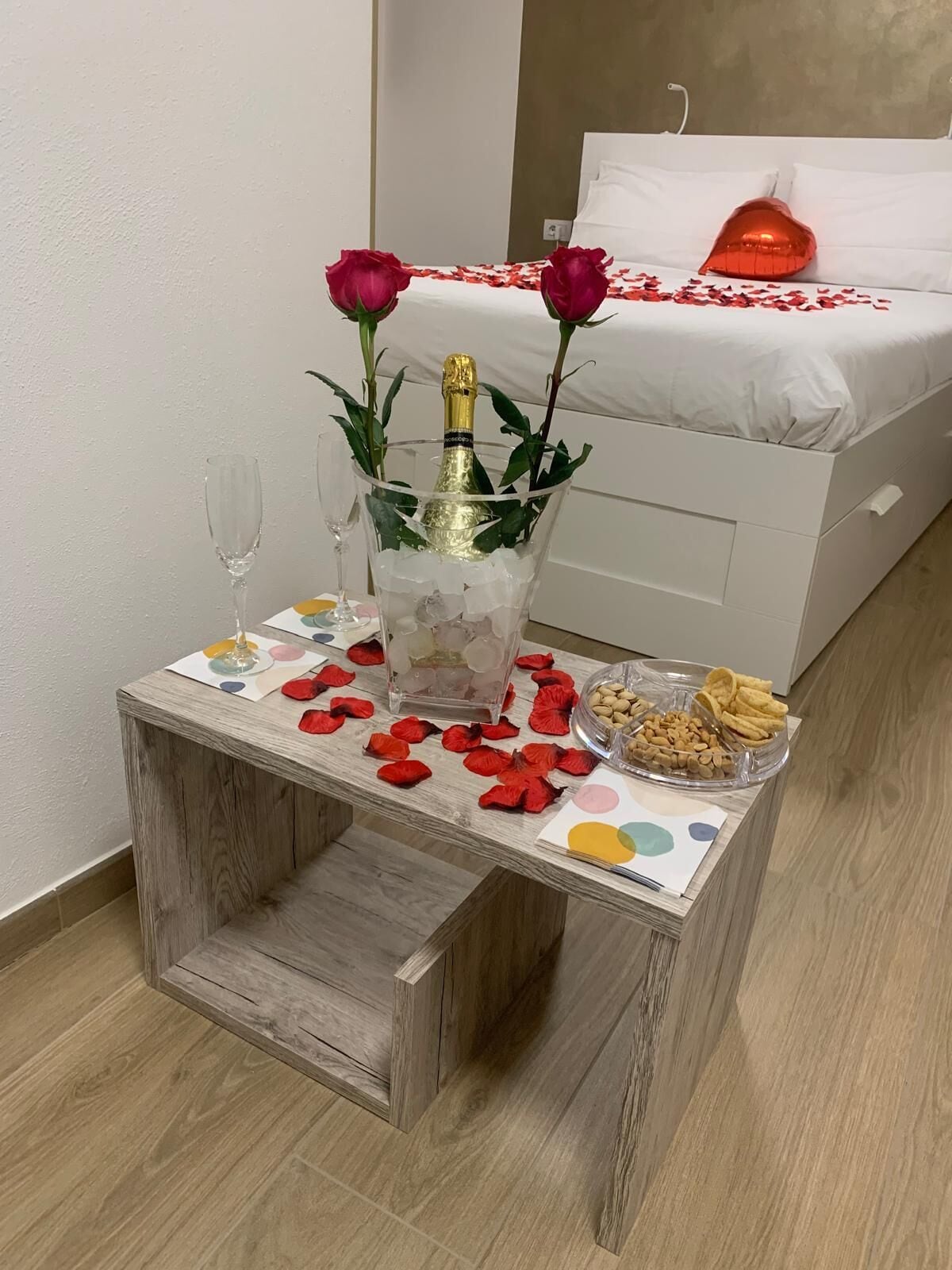 Et lille træbord med en champagneflaske, to roser og et champangneglas. Der ligger rosenblade spredt ud på bordet, samt en skål med nødder og kiks. Værelset har en seng med hvide sengetæpper og en rød ballon i hjerteform.