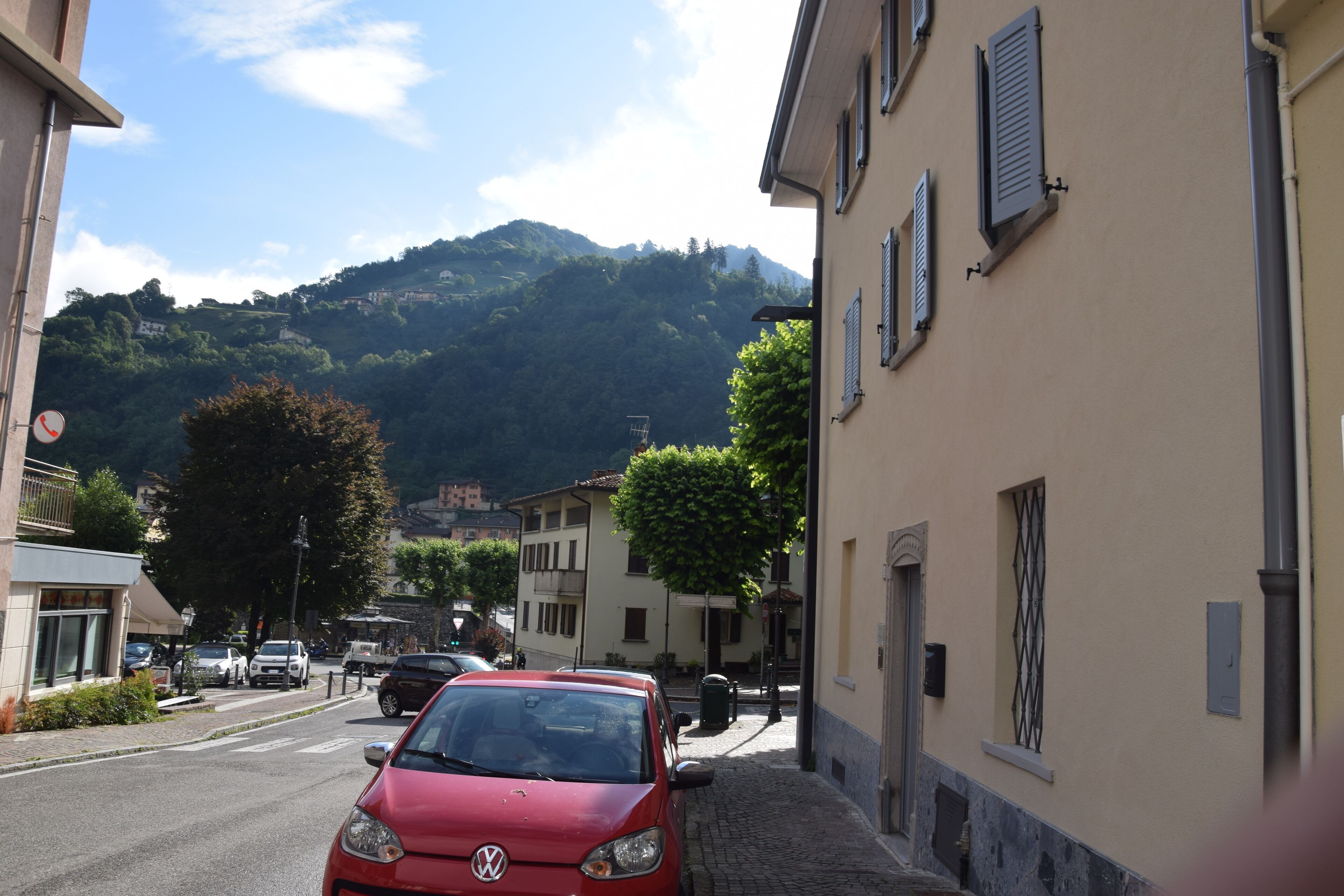A narrow street with parked cars and buildings on either side, leading to a hill with trees and houses in the background.
