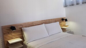 Quarto com cabeceira de madeira, cama com roupa de cama branca, duas mesas de cabeceira com abajures e uma janela com cortinas leves.