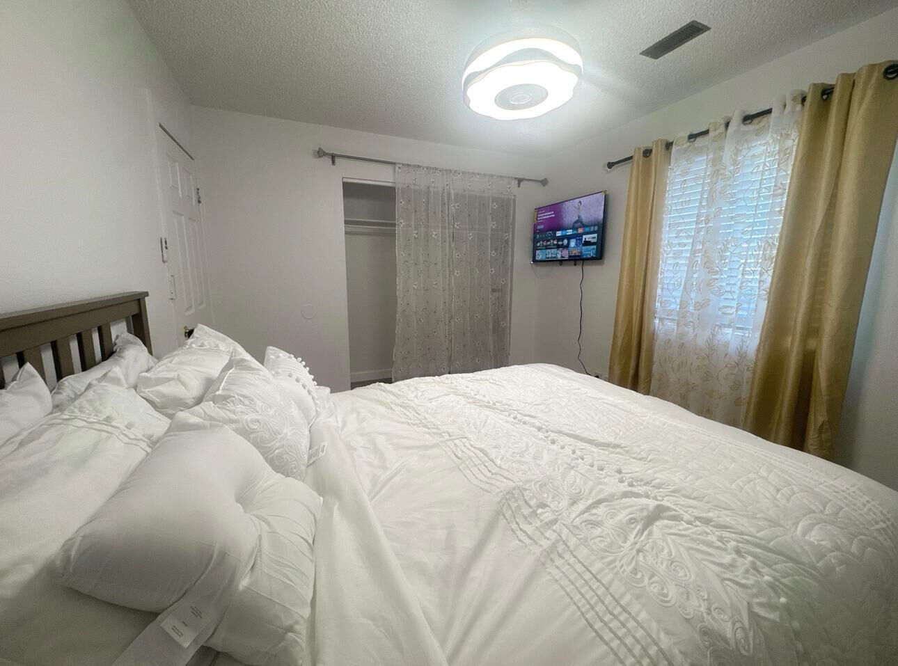 A neatly made bed with white linens and pillows, a television mounted on the wall, and a window with curtains.