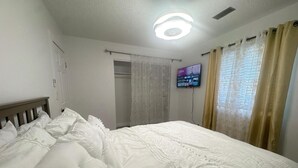 Un lit bien fait, avec du linge de lit blanc et des oreillers, un téléviseur fixé au mur et une fenêtre avec des rideaux.