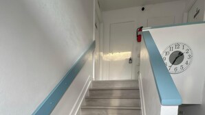 Un escalier avec des marches en bois clair et des murs blancs, menant à une porte. Sur le mur, à côté de la porte, on aperçoit une horloge et un extincteur.