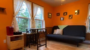 Ein gemütliches Zimmer mit Holzboden, einer schwarzen Couch, einem kleinen Tisch mit Hockern und einem Fenster mit Vorhängen.