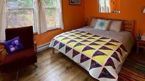 A bedroom with an orange wall, a wooden floor, a bed with a colorful quilt, a chair, and a window with curtains. - Trailside Cottage #2 (Victoria County)