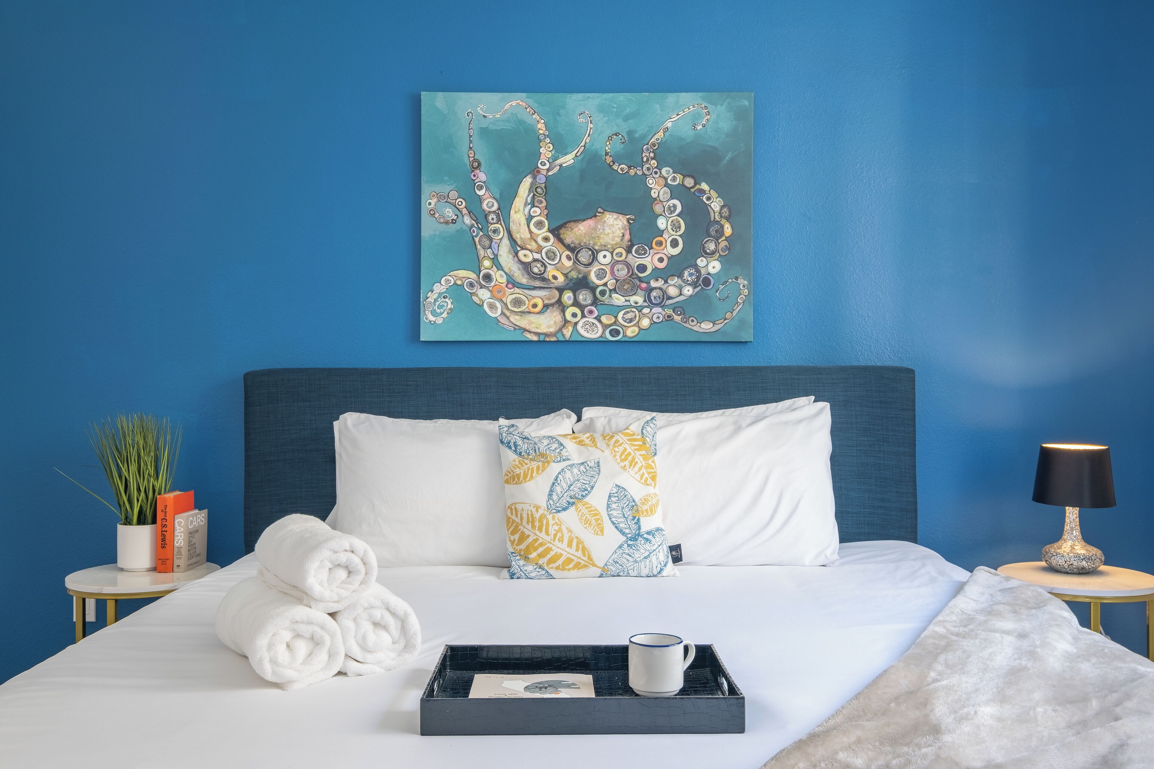 A neatly made bed with white linens, a blue wall, a painting of an octopus, and a small table with a lamp and a tray with a mug.