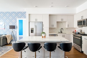 A modern kitchen with white cabinetry, a central island with bar stools, stainless steel appliances, and a patterned backsplash. - Beach Condo 234CB (Carlsbad)
