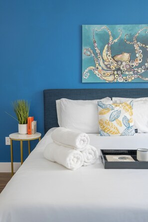 A neatly made bed with white linens, a blue wall, a painting of an octopus, a small round table with a plant, books, and a tray with a mug. - Beach Condo 234CB (Carlsbad)