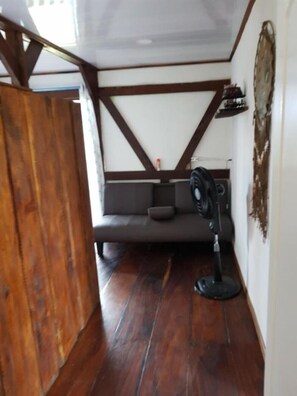 A room with a wooden floor, a grey sofa, a standing fan, and a wall-mounted shelf.