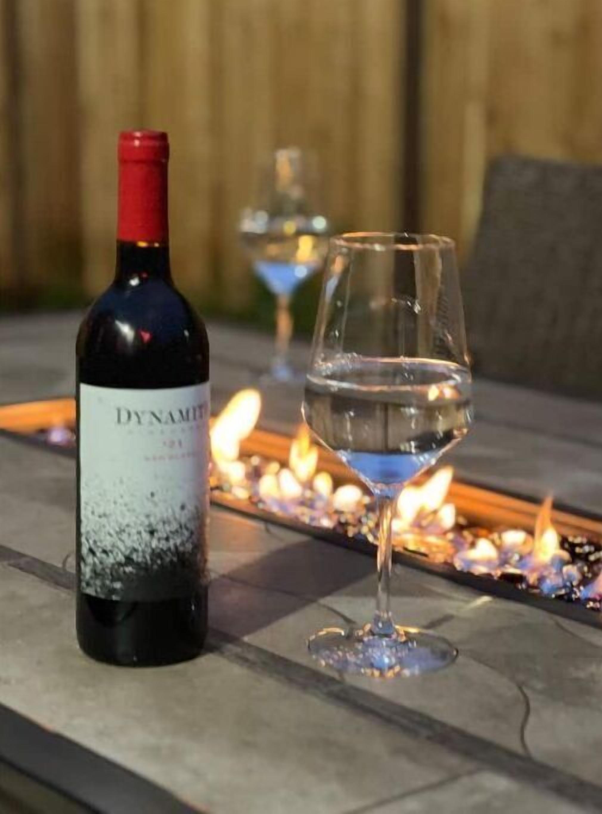 A bottle of wine and a glass on a table with a fire feature in the background.