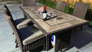 Een terras buiten met een vierkante tafel, rieten stoelen en een fles wijn.