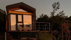 Una cabaña moderna con un amplio ventanal, terraza de madera y mobiliario exterior.