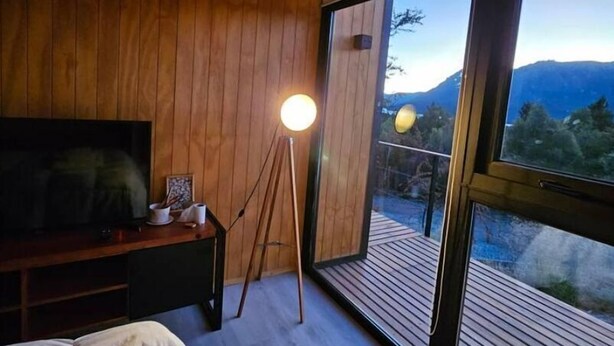 Un dormitorio con una cama grande, televisión, lámpara de pie y vistas a la montaña a través de amplios ventanales.