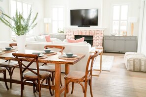 Ein Wohnzimmer mit weißem Sofa, einem Holzesstisch mit Stühlen, einem Fernseher an der Wand und einem Kamin aus Ziegelsteinen.