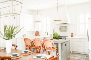 Una cocina luminosa con armarios blancos, un comedor con sillas de mimbre e iluminación colgante.