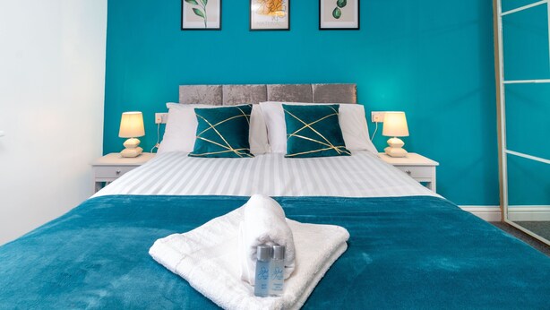 A bed with a teal bedspread, white pillows, and a white towel with toiletries. There are two bedside tables with lamps and a wall with three framed pictures. - Westlake Serviced Accommodation  (Peterborough)