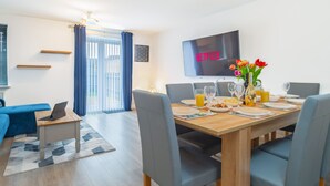 A modern dining area with a wooden table set for four, surrounded by grey upholstered chairs. A flat-screen TV mounted on the wall displays the word "NETFLIX". - Westlake Serviced Accommodation  (Peterborough)