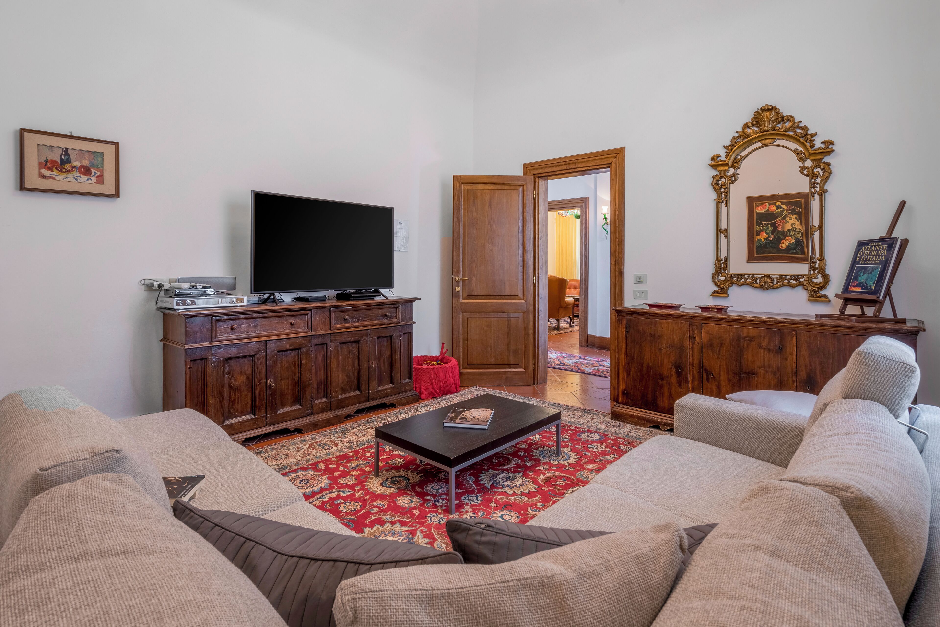 A living room with a large flat-screen TV, a wooden cabinet, a red ottoman, and a patterned rug.