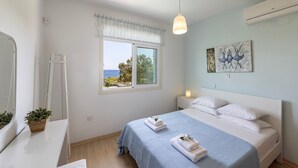 A bedroom with a bed, a bedside table with a plant, a window with a view, and a wall-mounted air conditioner.
