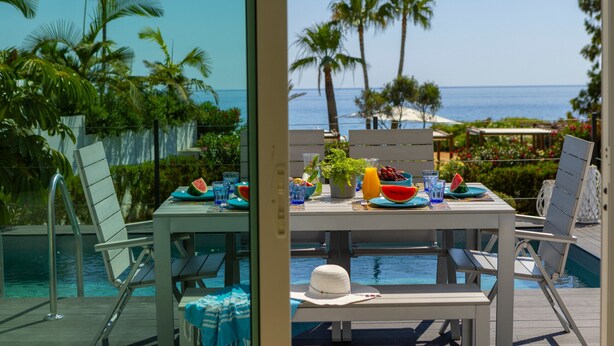 A poolside dining area with a table set for two, overlooking a pool and a beach with palm trees.