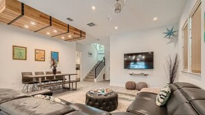 Uma sala de estar espaçosa com um amplo sofá de couro, uma área de jantar com mesa de madeira e uma TV fixada na parede.