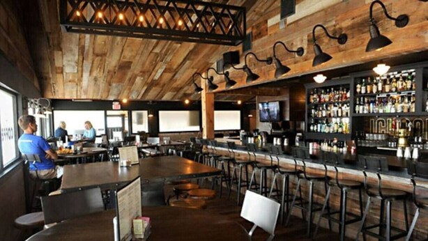 Um bar com teto de madeira e banquetas, um balcão com cardápio e uma tela de televisão.