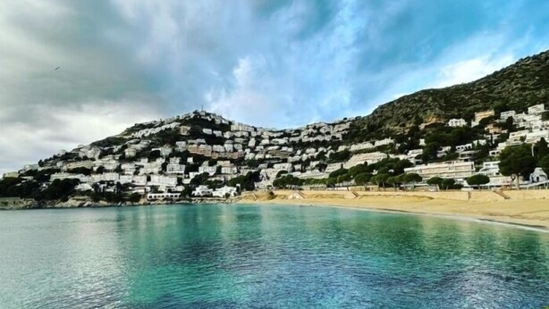 Une ville côtière avec des bâtiments blancs sur une colline, une plage et une eau d’un bleu limpide.