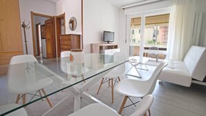 A modern living room with a glass dining table, white chairs, a sofa, a television, and a view of the outside. - AED Home   (Lavagna)
