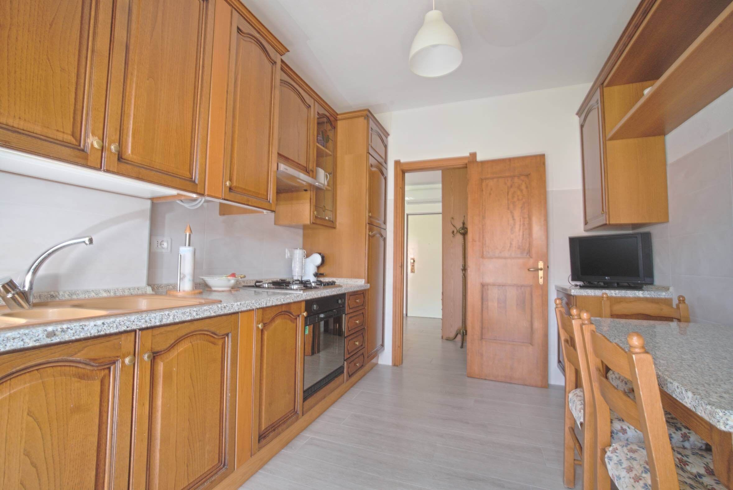 A kitchen with wooden cabinets, a granite countertop, a built-in oven, and a television on a wooden stand.