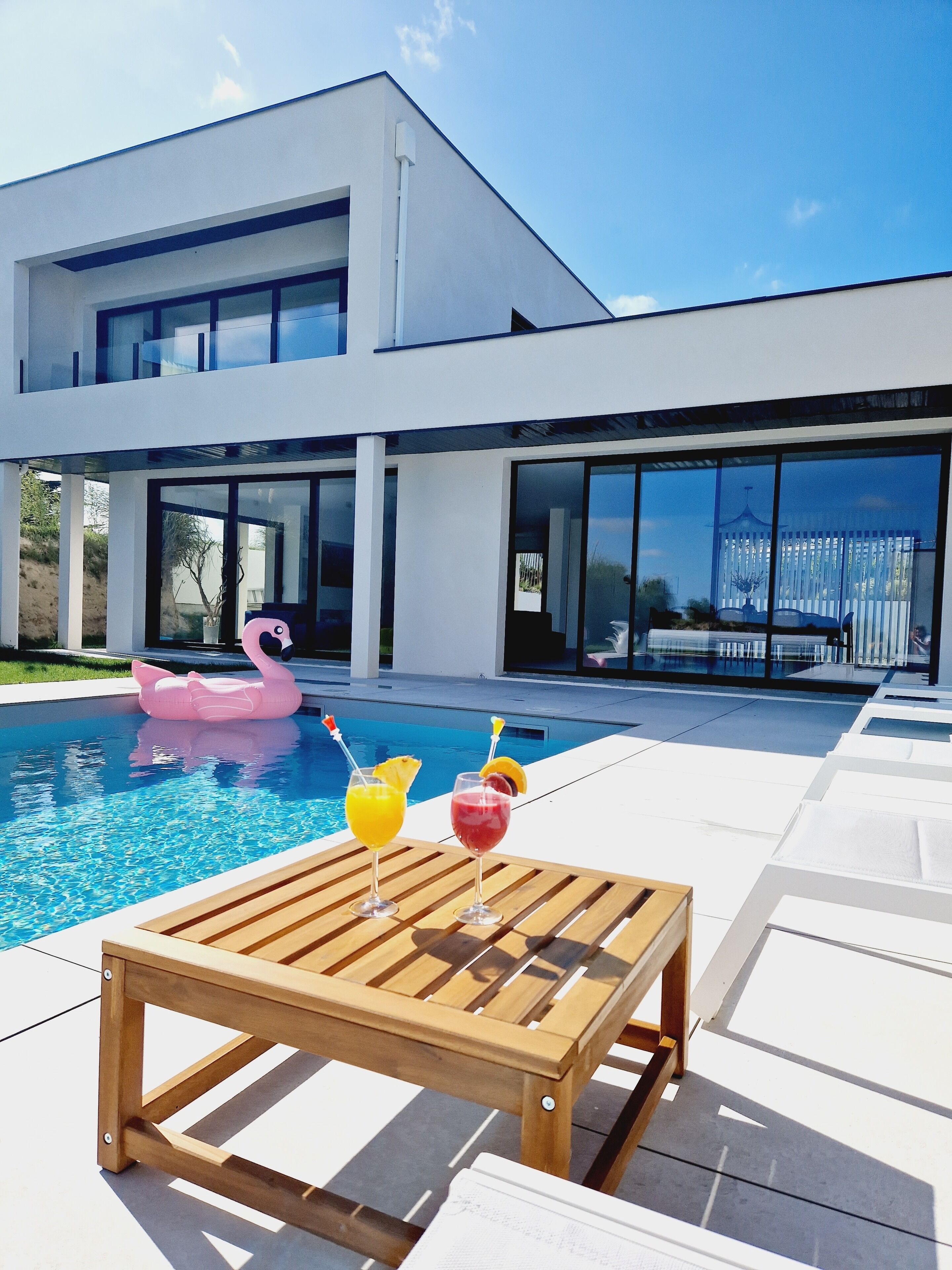 Una casa moderna con piscina, una mesa de madera con bebidas y un flotador con forma de flamenco rosa.