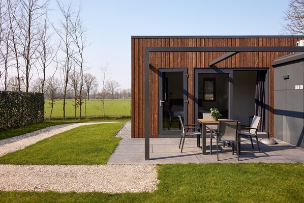 A modern building with a wooden exterior, a patio with a table and chairs, and a gravel path leading to it. - Hofparken Wiltershaar (Kotten)