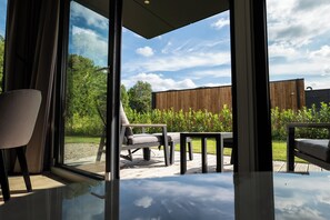 A modern outdoor seating area with a glass wall, wooden deck, and a view of a garden and trees. - Hofparken Wiltershaar (Kotten)