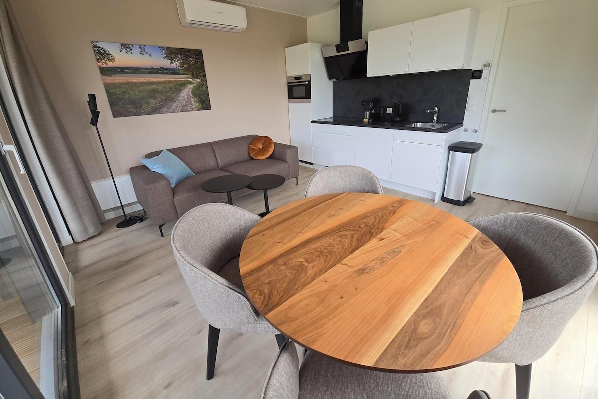 A modern living room with a round wooden table, grey armchairs, a sofa with cushions, a painting on the wall, and a kitchen area with white cabinets and a black countertop.