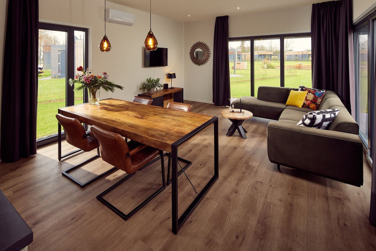 A modern living room with a wooden dining table, leather chairs, a sofa with colorful cushions, and large windows offering a view of the outdoors.