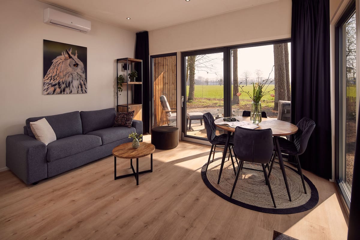 A modern living room with a sofa, coffee table, and dining area with chairs. There is a large window with a view of the outdoors.