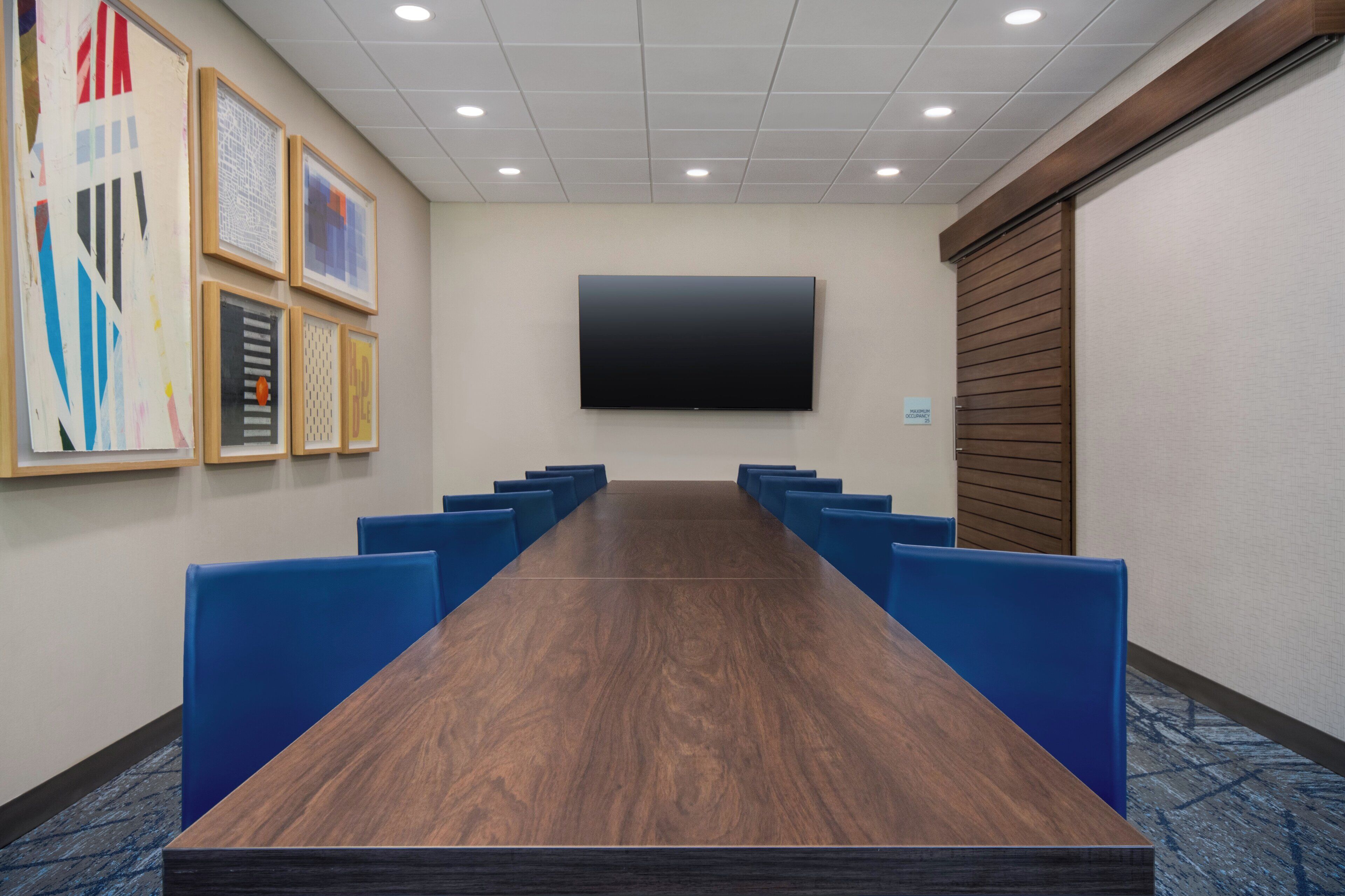 Una sala de conferències amb una taula llarga, cadires blaves, un televisor de pantalla plana i quadres a les parets.