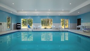 A swimming pool with clear blue water, surrounded by white chairs and a table, with large windows offering a view of trees. - Holiday Inn Express & Suites Muskegon - Grand Haven by IHG (Muskegon)