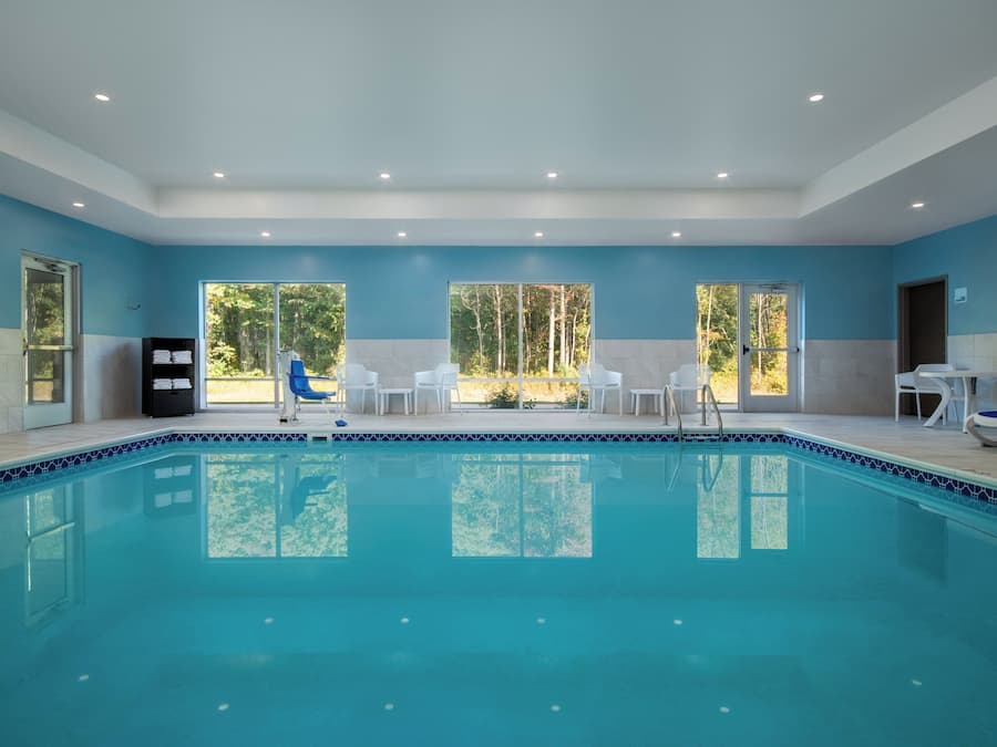 A swimming pool with clear blue water, surrounded by white chairs and a table, with large windows offering a view of trees.