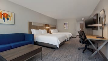Une chambre d’hôtel avec un canapé bleu, deux lits avec du linge de lit blanc, un bureau avec une chaise, un téléviseur à écran plat et des œuvres d’art accrochées aux murs.