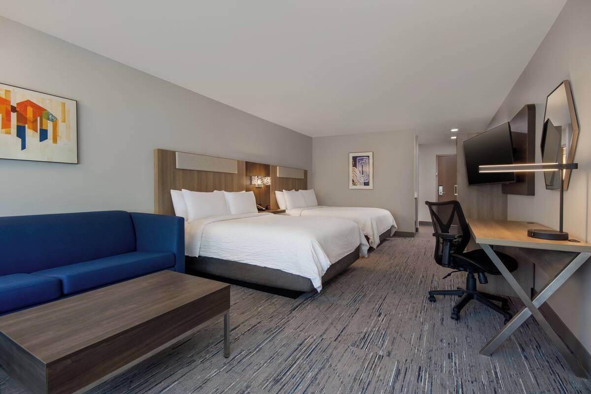 A hotel room with a blue sofa, two beds with white bedding, a desk with a chair, a flat-screen TV, and artwork on the walls.