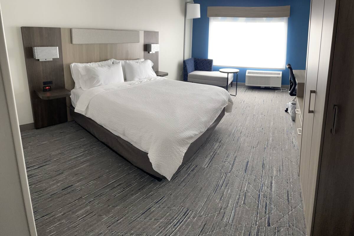 A hotel room with a large bed, a bedside table with a lamp, a small table with a chair, and a window with a view.