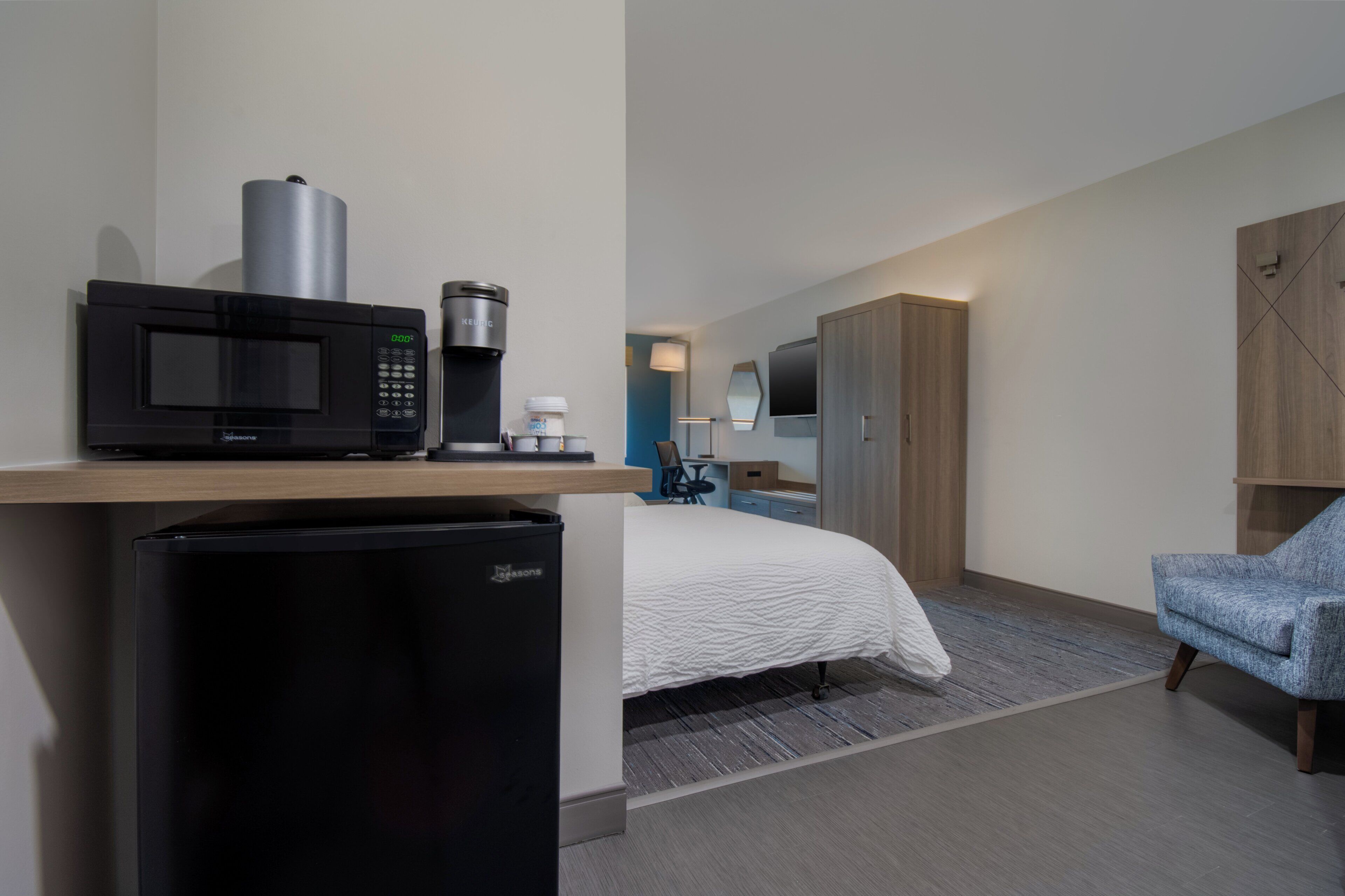 A hotel room with a bed, a desk with a microwave, a refrigerator, a chair, and a wardrobe.