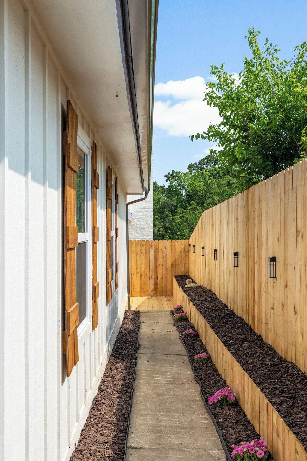 A narrow pathway leading to a wooden fence with small windows, surrounded by a white building and greenery.